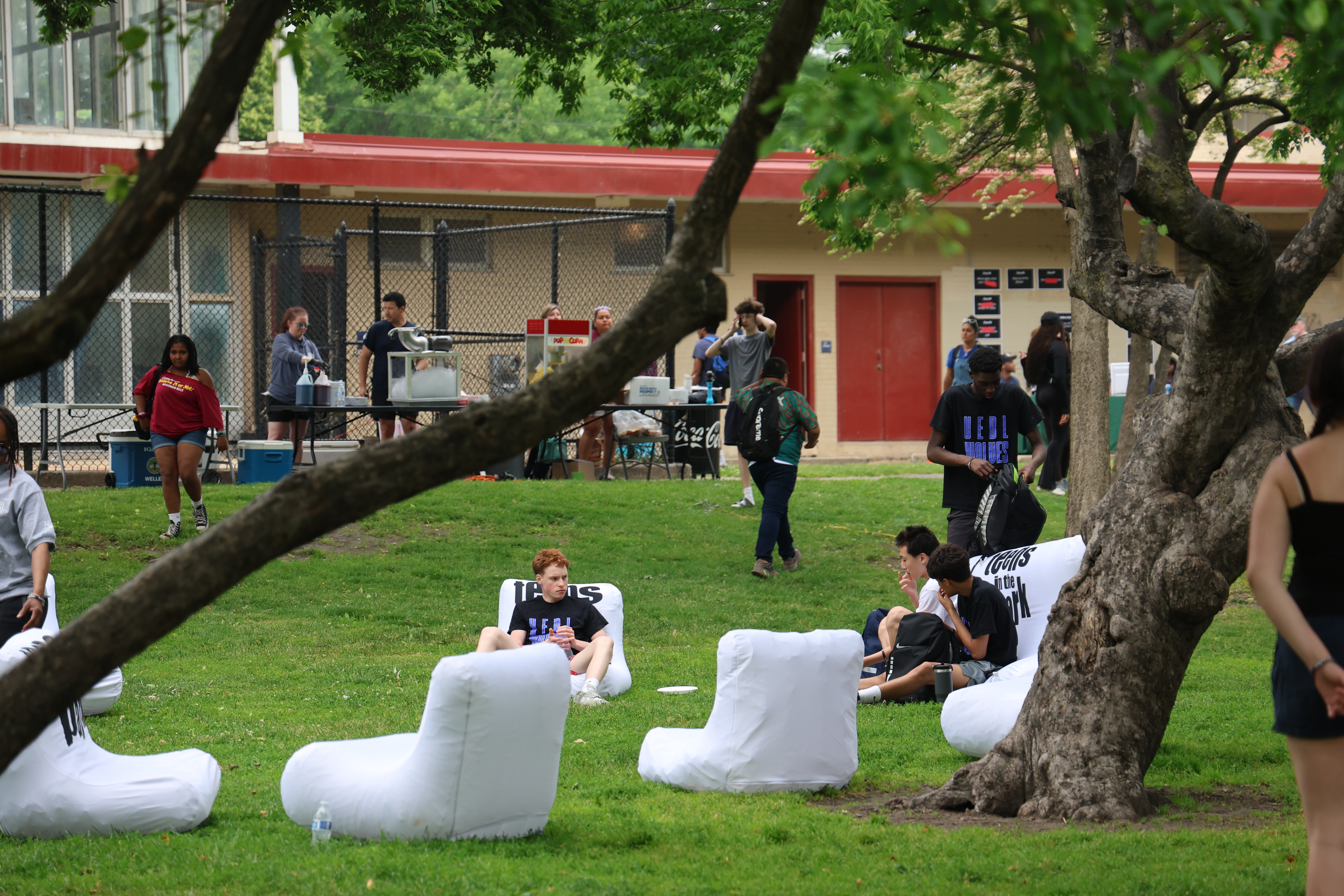Students relax in inflatable chairs on a grassy area during a school event. A popcorn machine and other refreshments are visible in the background.